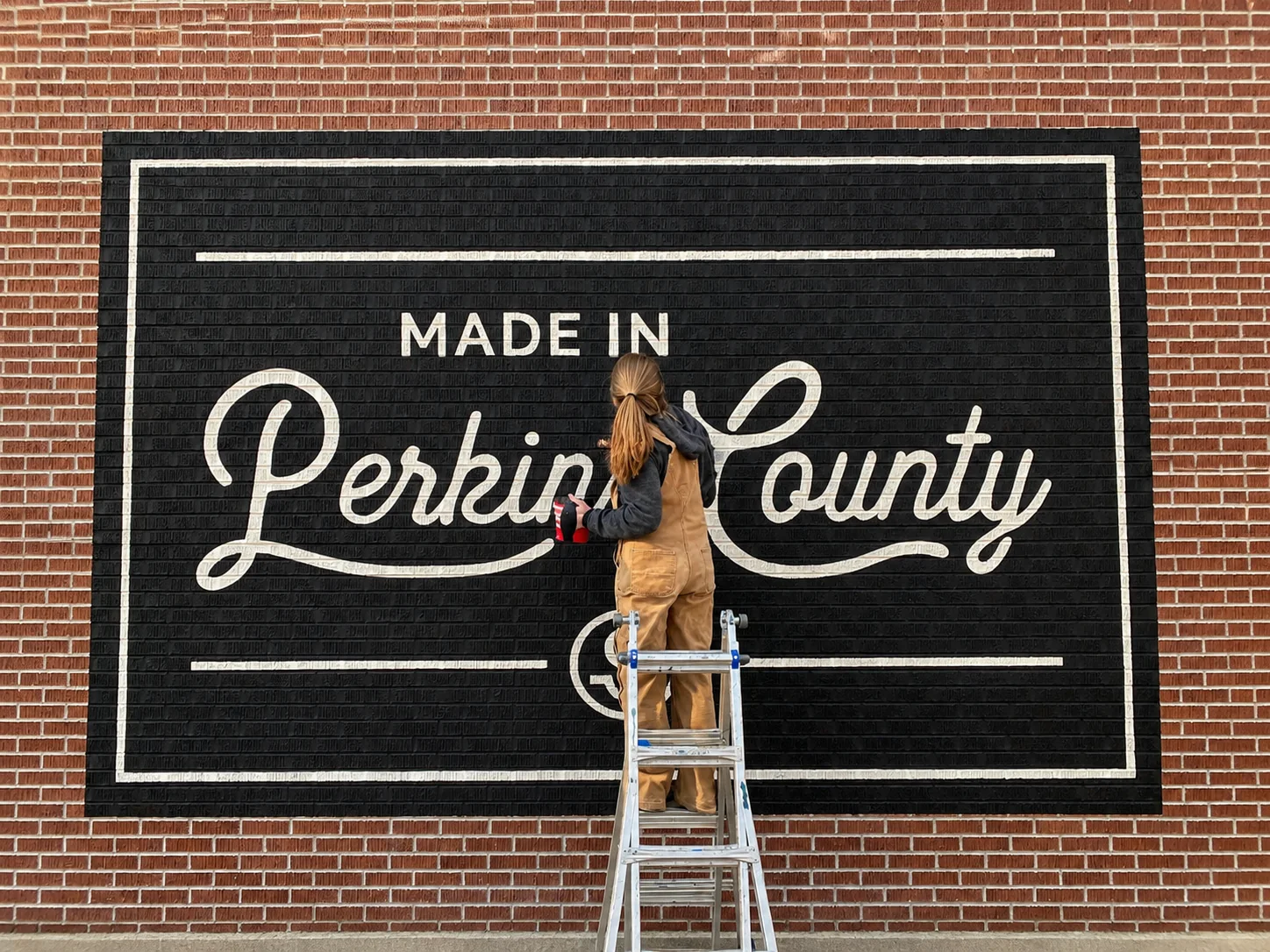Erin Larwick painting white script on the Made in Perkins County mural from a ladder